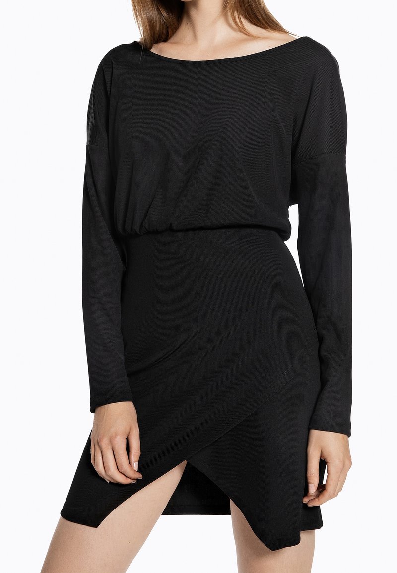 Femme portant une robe noire à manches longues avec un large décolleté et une jupe portefeuille asymétrique dévoilant la cuisse.