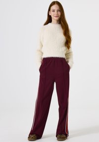 Suéter blanco de felpa con cuello redondo, combinado con pantalones burdeos que tienen rayas laterales blancas y rojas. Llevado con zapatos de estampado animal.