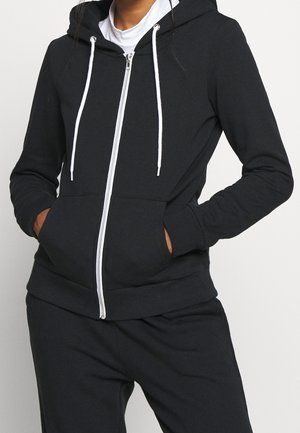 Person trägt schwarzen Kapuzenpullover mit Reißverschluss und weißen Kordeln sowie schwarze Hose, Hände in den Taschen des Kapuzenpullovers, vor einfarbigem Hintergrund.