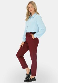 Kobieta pozująca w jasnoniebieskiej bluzce z długim rękawem, bordowych rurkach z elastycznym paskiem w talii oraz czarnych lakierowanych mokasynach na białym tle.