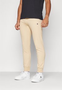 Beige stretch sweatpants with elastic cuffs, featuring a small logo on the left thigh. Paired with white athletic shoes and a black shirt.