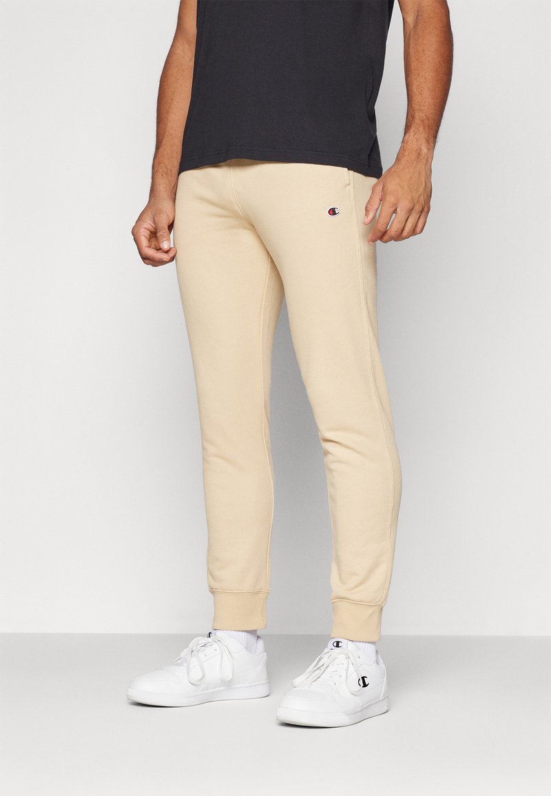 Beige stretch sweatpants with elastic cuffs, featuring a small logo on the left thigh. Paired with white athletic shoes and a black shirt.