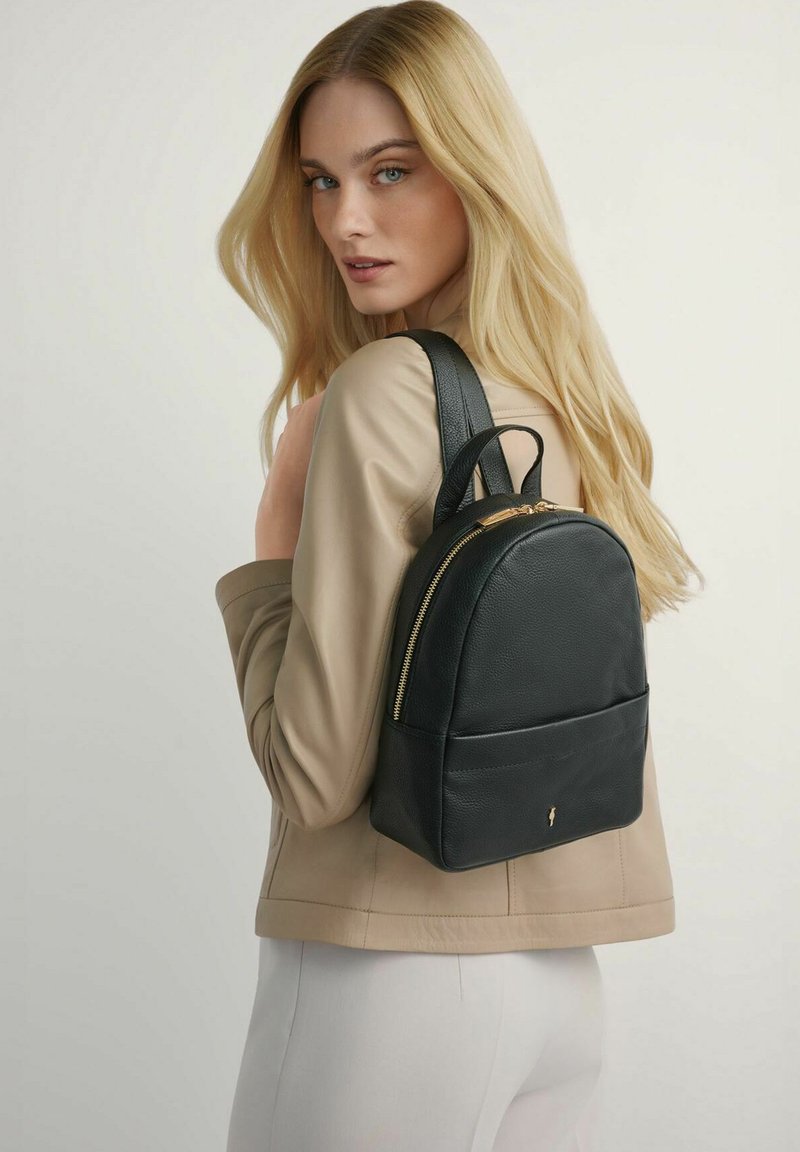 Blondynka w beżowej kurtce, niosąca na ramieniu czarny skórzany plecak ze złotym zamkiem błyskawicznym, spoglądająca za siebie.