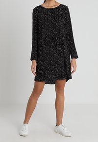 Nainen, joka on pukeutunut mustaan pitkähiharaiseen mekkoon, jossa on pieniä vaaleanpunaisia ja valkoisia kuvioita, yhdistettynä valkoisiin sneakereihin, seisoo yksinkertaista taustaa vasten.