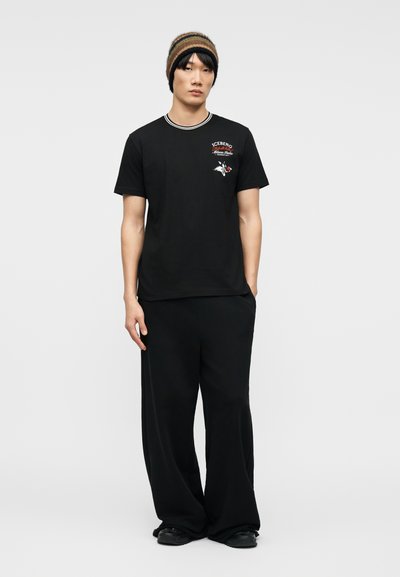 Νεαρός άνδρας που φοράει μαύρο t-shirt με γραφικό σχέδιο, φαρδιά μαύρη παντελόνια, μαύρα παπούτσια και καφέ ριγέ πλεκτό σκουφάκι, στέκεται μπροστά σε απλό φόντο.