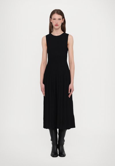 Robe noire sans manches avec un corsage ajusté et une jupe plissée, faite en tissu côtelé. Assortie à des bottes noires hauteur genou. Design simple.