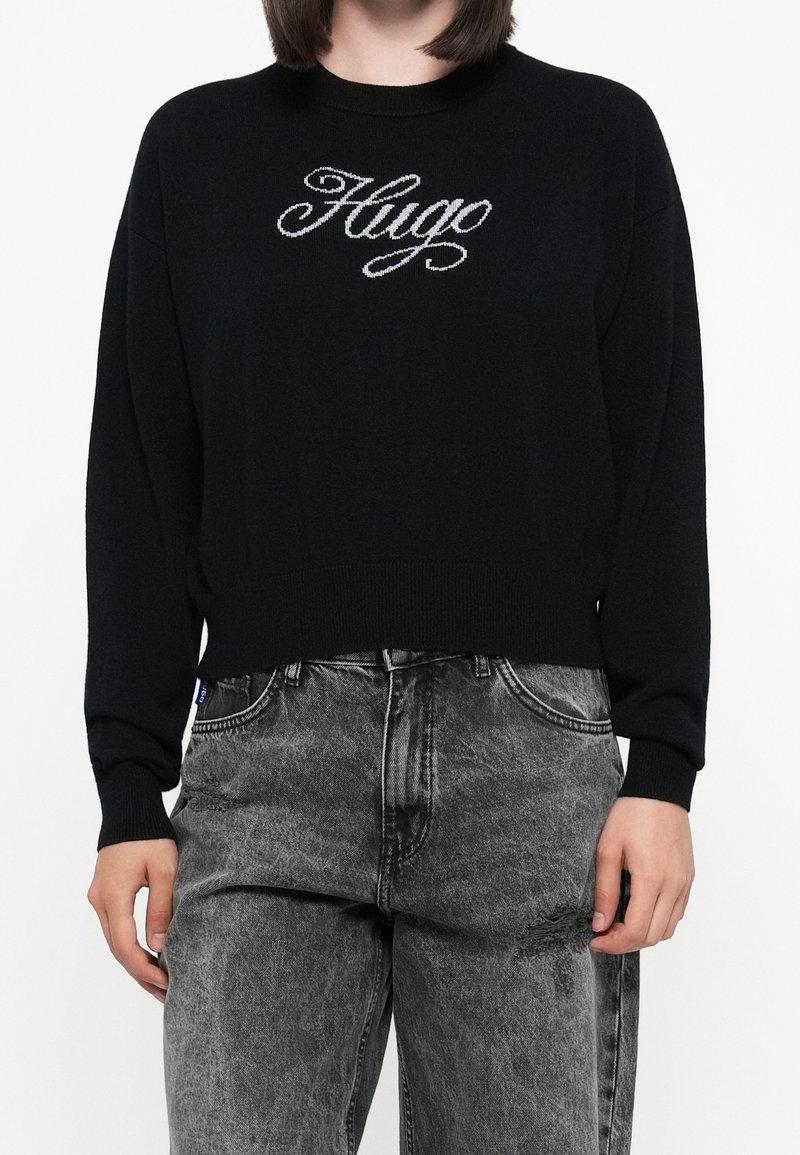 Personne portant un pull noir avec « Hugo » en écriture blanche et un jean noir délavé taille haute avec une petite déchirure sur la cuisse.