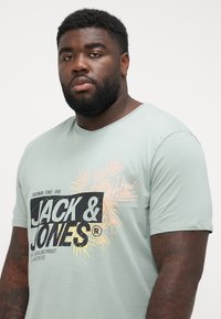 Light green cotton t-shirt featuring a black and yellow graphic print with "JACK & JONES" text and palm leaf design. Short sleeves, round neck.