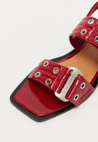 Red patent leather sandal with a square toe. Features two wide straps with grommets and a metal buckle displaying the brand name. Black sole.