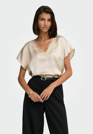 Femme aux cheveux courts et foncés portant un chemisier en satin crème avec bordure en dentelle et un pantalon noir taille haute, debout les mains jointes.
