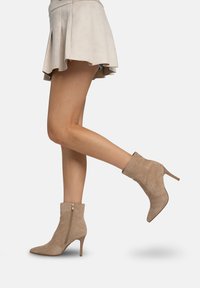 Botines beige de ante con punta afilada y tacón de aguja fino. Presentan una cremallera lateral y textura lisa.