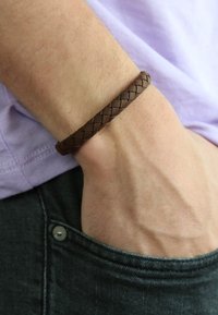 Brown braided leather bracelet on a wrist, set against a light purple shirt and denim. Features a textured, woven design.