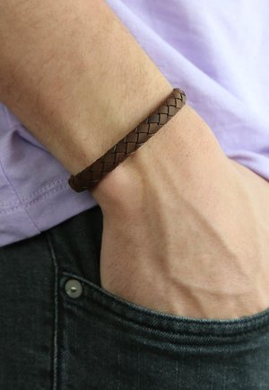 Brown braided leather bracelet on a wrist, set against a light purple shirt and denim. Features a textured, woven design.