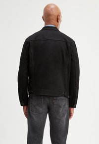 Black denim jacket with a classic cut, featuring side button closures and a smooth finish. Paired with faded gray jeans.
