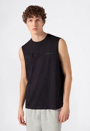 Joven con cabello rizado vistiendo una camiseta negra sin mangas de Champion y pantalones deportivos gris claro sobre un fondo liso.