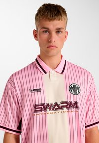 Pink og cremefarvet polo med striber og knaplukning ved kraven, med et logo på brystet og sorte detaljer på kraven og ærmerne.