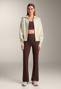 Beige sweater met rits en hoge kraag, gecombineerd met een bruin cropped topje en wijde broek. Lichtgekleurde sneakers maken de outfit af.