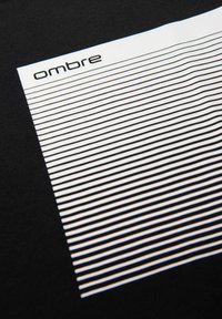 Černé tričko s vodorovným bílým pruhovaným vzorem různých šířek, označené nápisem "ombre" v elegantním písmu na vrchu.
