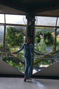 Femeie în costum albastru stă desculță sprijinită de o balustradă în fața unor ferestre mari, cu verdeață în exterior și un ventilator de tavan deasupra.