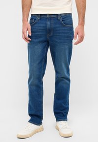 Jeans de mezclilla azul con corte recto, diseño de cinco bolsillos, costuras en contraste y textura desgastada, combinados con zapatillas blancas.