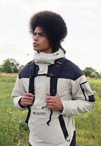 Lett, beige og marineblå jakke med høy krage, frontlomme og justerbare stropper. Har glidelåser og elastiske mansjetter.