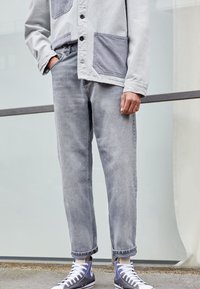 Chaqueta color gris claro con botones y dos grandes bolsillos tipo parche, combinada con jeans de mezclilla gris claro y zapatillas altas azules.