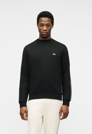 Jeune homme portant un sweat-shirt noir avec un logo sur la poitrine et un détail rayé sur les manches, associé à un pantalon clair.