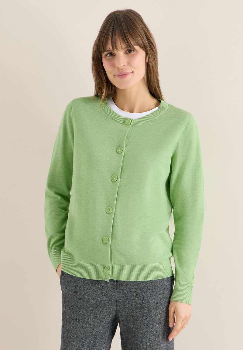 Femme portant un cardigan vert clair boutonné sur une chemise blanche et un pantalon gris foncé, debout contre un mur beige uni.