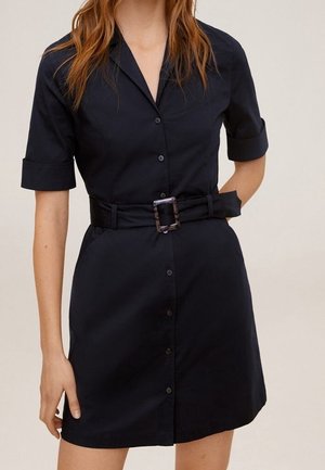 Femme portant une robe noire à manches courtes boutonnée, avec un col et une large ceinture avec une grande boucle carrée.