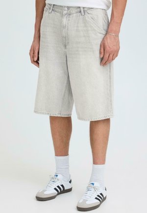 Homme portant un short en denim gris clair atteignant les genoux, des chaussettes blanches montantes et des baskets blanches avec des rayures noires, debout devant un fond uni.
