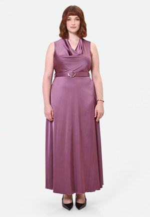 Donna con capelli rossi fino alle spalle che indossa un vestito senza maniche in raso malva con cintura abbinata e tacchi neri a punta, in piedi davanti a uno sfondo semplice.
