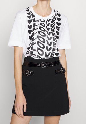Persoon draagt een wit T-shirt met zwarte tekst "LOVE" en hartjes, ingestopt in een zwarte rok met drie zwarte riemen van lakleer met gespen.