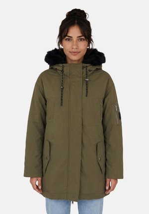 Donna che indossa un parka verde oliva con cappuccio bordato di pelliccia nera, tasche frontali e tasca con zip sul braccio, in piedi davanti a uno sfondo semplice.
