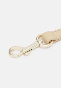 Mousqueton métallique doré attaché à une sangle en cuir beige avec des coutures visibles sur fond blanc.