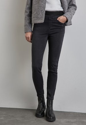 Person iført sorte skinny jeans, sorte ankelstøvler, grå tekstureret jakke og hvid top, stående mod en ensfarvet grå væg.