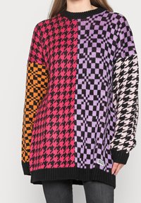 Bunter gestrickter Pullover mit einem markanten Schachbrettmuster in Pink, Orange, Schwarz und Lila, ausgestattet mit einem schwarzen, gerippten Rundhalsausschnitt und Bündchen.