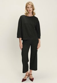 Mujer con cabello rubio que lleva una blusa negra de manga larga con textura y pantalones de pierna ancha a juego, además de tacones puntiagudos burdeos.