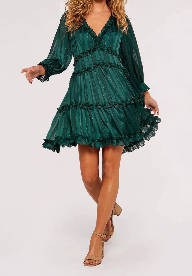 Femme portant une robe longue à manches longues verte foncée avec volants et manches transparentes, ainsi que des sandales à talons hauts beige, debout avec une jambe croisée.