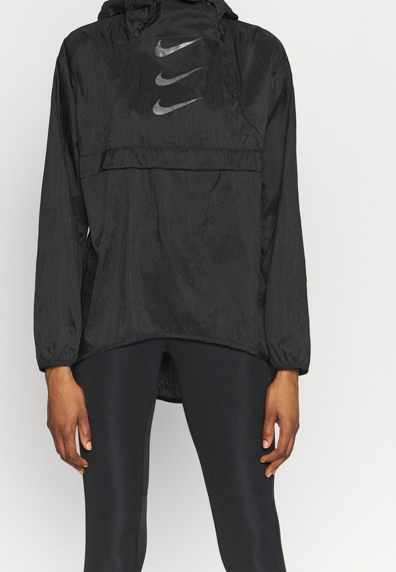 Zwarte Nike windbreaker met een capuchon, lange mouwen en een reflecterend logo op de borst. Het materiaal heeft een gestructureerde afwerking en een losse pasvorm.