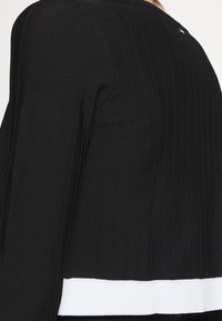 Jersey de punto negro acanalado, cropped, que presenta mangas largas, un escote redondeado y un detalle de dobladillo blanco en la parte inferior. Sutil acento de logo en la parte posterior.