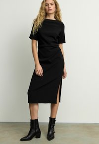 Robe midi noire à manches courtes, avec une fente sur le côté et une silhouette ajustée. Portée avec des bottines noires et une texture lisse et mate.
