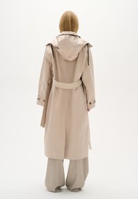 Béžový voděodolný trench coat s kapucí, páskem v oblasti pasu a knoflíkovými manžetami. Hladká textura, délka pod kolena a rozparky vzadu.