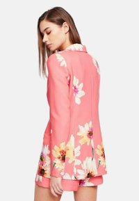 Blazer rosa con motivo floreale in giallo e bianco. Design sartoriale con colletto, maniche lunghe e spacco sul retro. Tessuto dalla consistenza liscia.