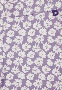 Lila tyg med vita blommönster i varierande former och storlekar. Inkluderar en liten lila etikett med en vit stjärndetalj.