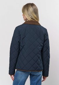 Vrouw met de rug naar de camera, gekleed in een marineblauw doorgestikt jack met een bruine kraag en bies, gecombineerd met een blauwe spijkerbroek, tegen een effen achtergrond.