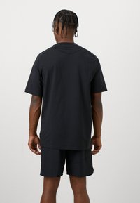 T-shirt in cotone nero con maniche corte, taglio rilassato e orlo curvo. Modello abbinato a pantaloni corti neri, mostrando un design semplice e minimale.