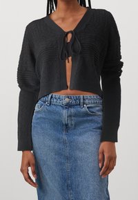 Černý pletený kardigan s krátkým střihem, s texturovanými vzory a zavazováním v přední části, v kombinaci s modrou džínovou sukní.