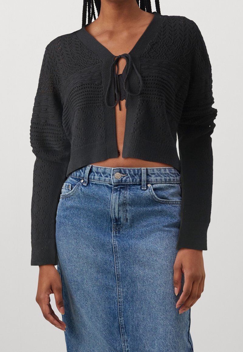 Černý pletený kardigan s krátkým střihem, s texturovanými vzory a zavazováním v přední části, v kombinaci s modrou džínovou sukní.
