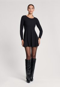 Abito nero a maniche lunghe con coste, dotato di gonna a ruota, abbinato a stivali in pelle nera alti fino al ginocchio. Tessuto liscio e design aderente.