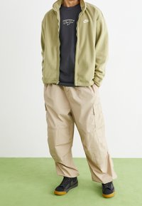 Grön fleecejacka, svart logot-shirt, beige cargobyxor och svarta sneakers med orange sulor. Slät textur; avslappnad passform; praktisk design.